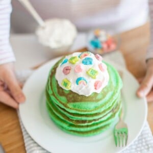 A person holds a plate of green pancakes covered with whipped cream and colorful cereal pieces. A fork rests on the plate. A small bowl of cereal and a container of whipped cream are in the background on a wooden surface.