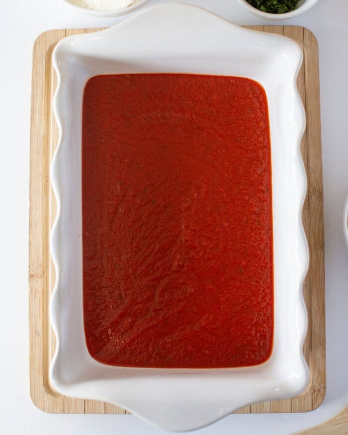 A white rectangular baking dish filled with smooth red tomato sauce on a wooden board. Small portions of ingredients like flour and herbs are partially visible around the dish, suggesting the preparation of a recipe.