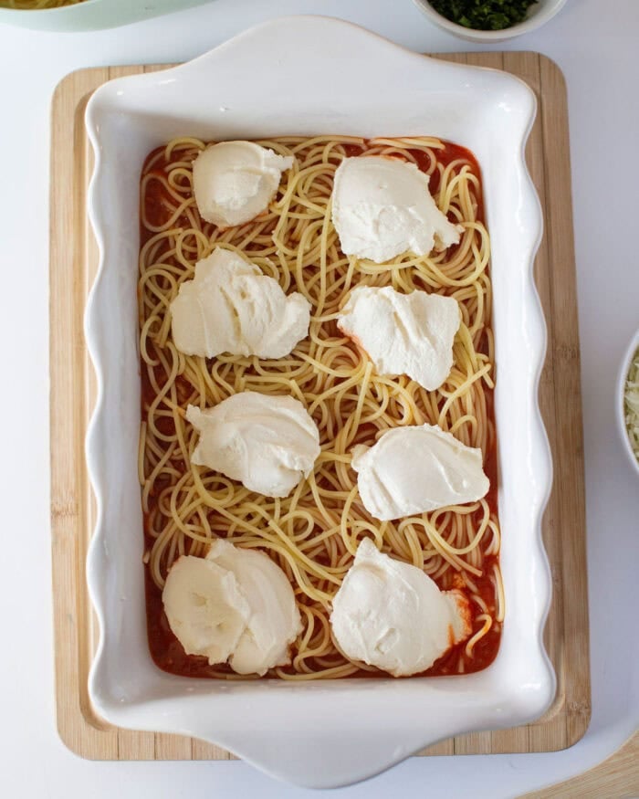 A casserole dish with a layer of cooked spaghetti spread over a red sauce, topped with spoonfuls of ricotta cheese. The dish is placed on a wooden cutting board.