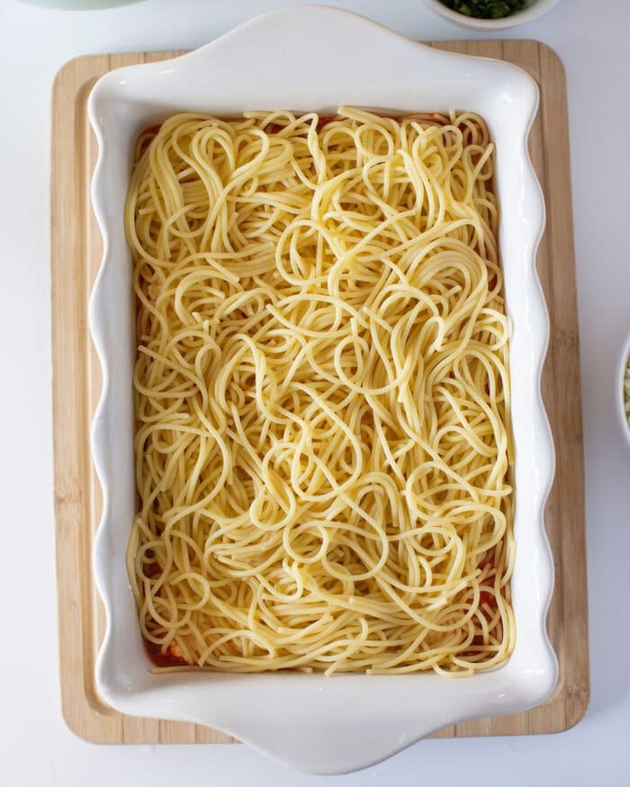 A rectangular white ceramic baking dish filled with cooked spaghetti, placed on a wooden board. The pasta is neatly arranged and ready for further preparation or serving.