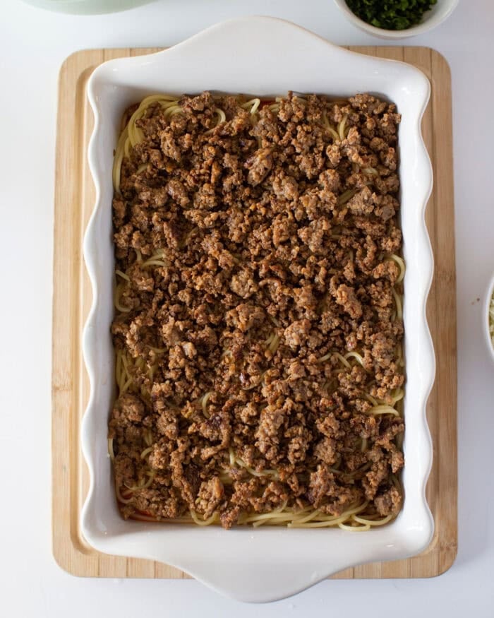 A white baking dish filled with cooked ground meat layered over spaghetti sits on a wooden cutting board. Small bowls with green herbs are visible in the background.
