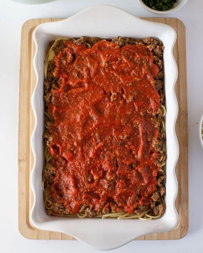 A white rectangular baking dish filled with cooked spaghetti topped with ground beef and rich red tomato sauce, placed on a wooden cutting board. Small bowls with chopped herbs are visible at the top.