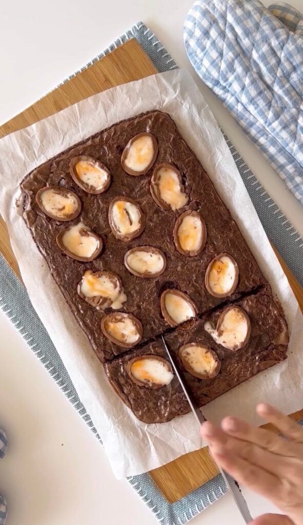 Slicing with a knife baked cadbruy egg brownies on a wood cutting board.