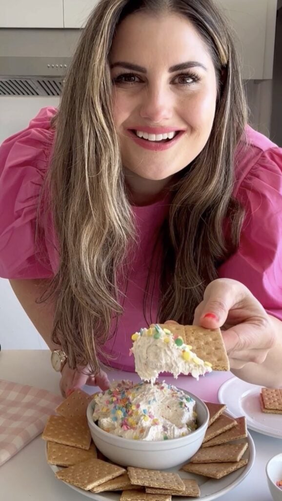 Melissa wearing a pink shirt holding a graham cracker dipped in Funfetti Dip.