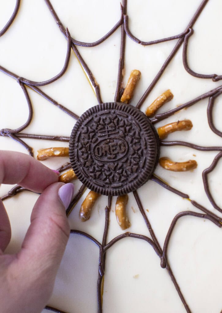 A hand is arranging a chocolate cookie with pretzel sticks to resemble a spider. The setup is placed on a web design made of chocolate icing on a white surface.