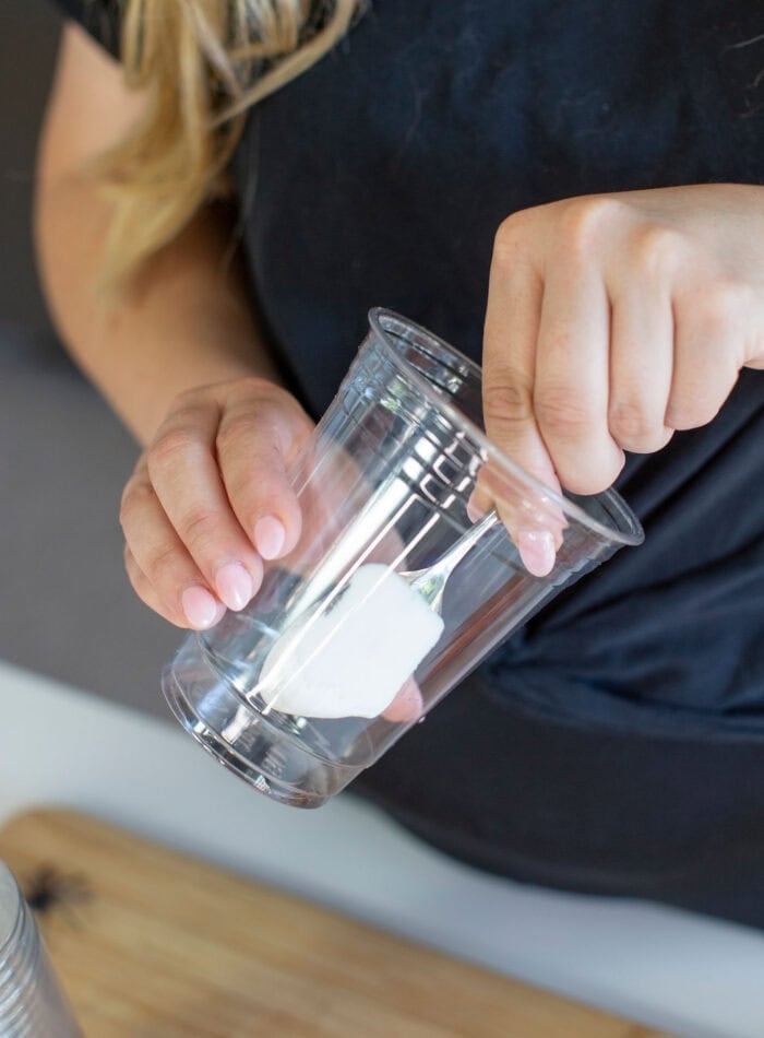 Person holding a clear cup with a spoonful of white powder inside, possibly preparing a mixture. They are wearing a dark shirt, and the background includes a wooden surface.
