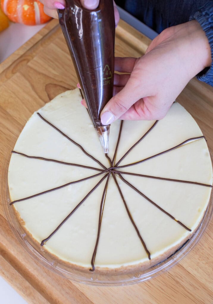 A person is piping dark chocolate lines on a cheesecake, dividing it into equal slices. The cheesecake is on a wooden surface, and theres a pumpkin partially visible in the background.