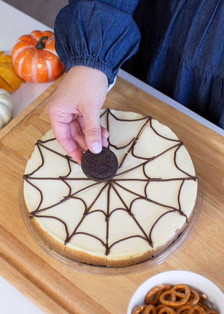 A person is placing a chocolate cookie on a cheesecake with a chocolate spiderweb design. The cake sits on a wooden board. Small pumpkins and pretzels are nearby.
