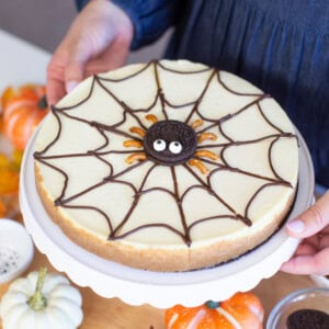 Person holding a cheesecake decorated with a chocolate spider web and a cookie spider in the center. The cake is on a white plate. Nearby are decorative pumpkins and autumn leaves on a wooden board.