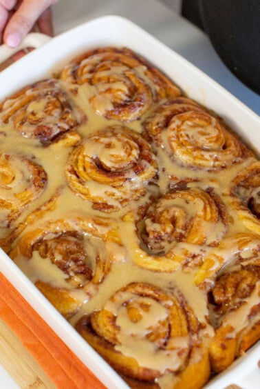 A close-up of freshly baked cinnamon rolls in a rectangular white dish, topped with a creamy glaze. A hand holds one side of the dish. The rolls are tightly packed and glistening with the glaze.