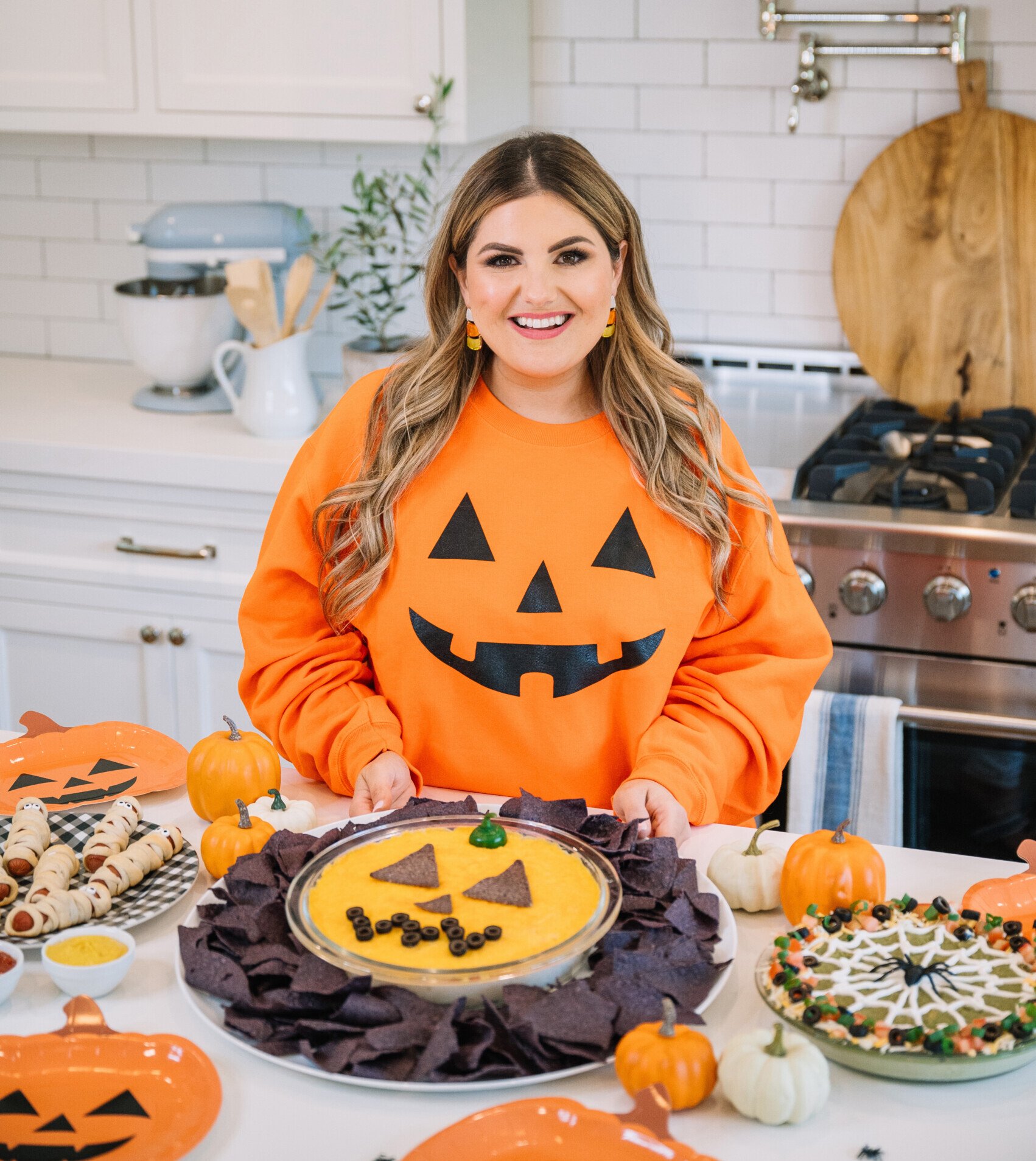 A woman wearing an orange pumpkin-face sweater stands in a bright kitchen, smiling behind a table with Halloween-themed snacks, including a pumpkin-shaped dip and other festive treats.