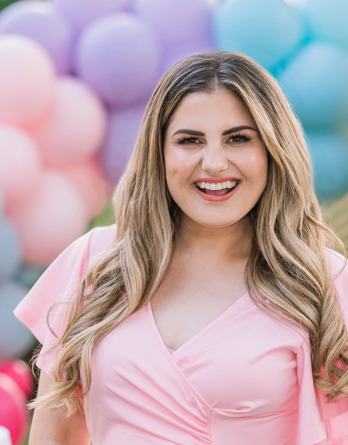 A woman with long, wavy blonde hair in a light pink dress smiles in front of pastel-colored balloons in pink, purple, and blue.