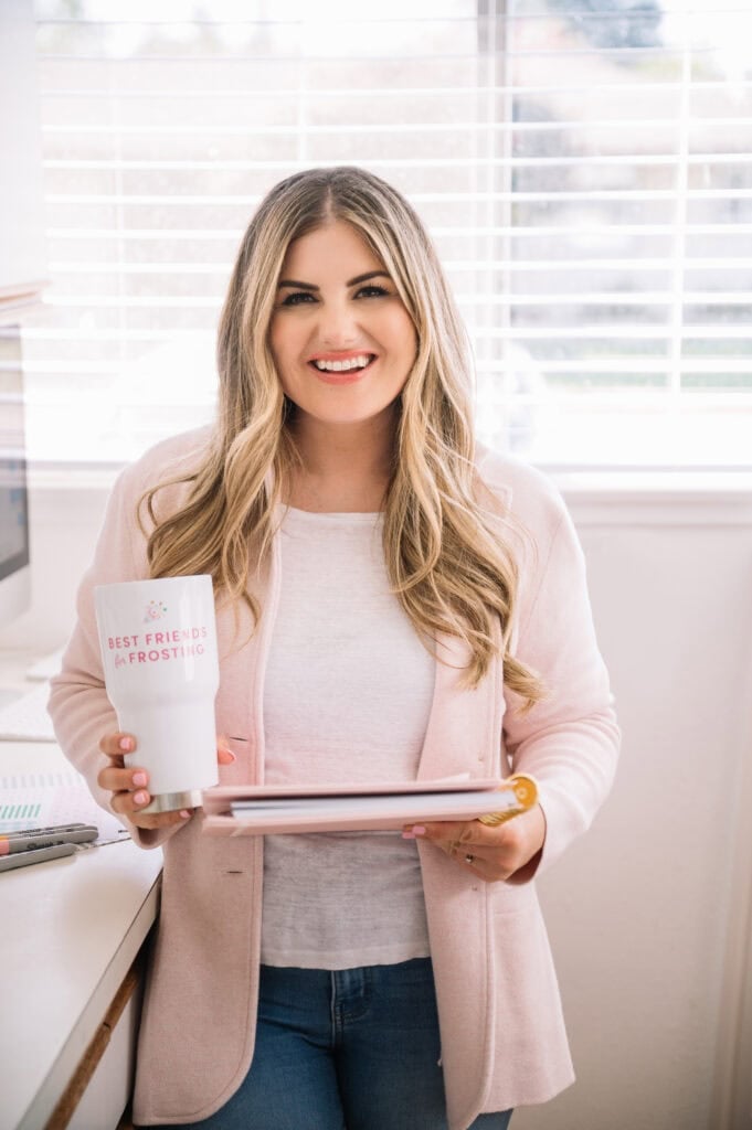 A woman with long blonde hair is smiling while holding a tumbler and a pink notebook. Shes dressed in a light pink blazer over a white top, standing by a window with blinds. The tumbler reads Best Friends & Frosting.