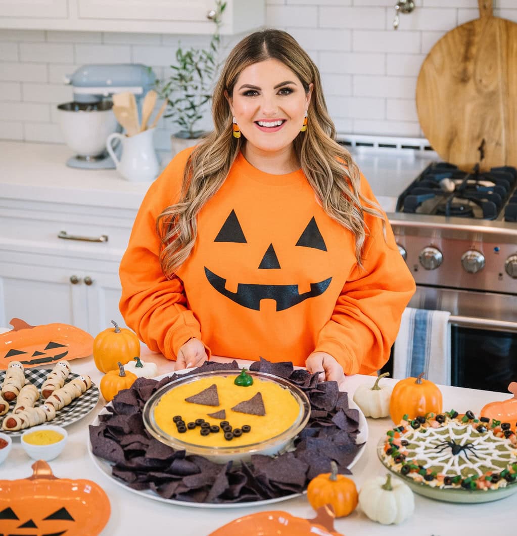 A woman in a pumpkin face sweater stands in a white kitchen, surrounded by Halloween-themed food: a pumpkin-shaped cheese dip with tortilla chips, mummy hot dogs, and a spider web pie. Small pumpkins decorate the counter.
