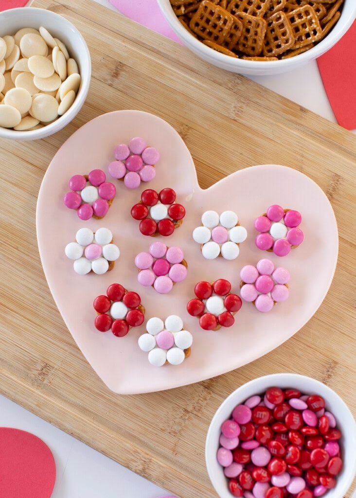 Close-up of M&M pretzel flowers on a pink heart-shaped plate. The flowers are made with red, pink, and white M&M's arranged around pretzel snaps.