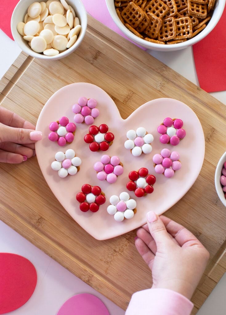 Two hands holding M&M pretzel flowers on a pink heart-shaped plate. The flowers are made with red, pink, and white M&M's arranged around pretzel snaps.