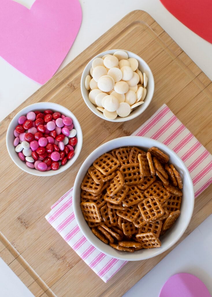 Valentine's pretzel bite ingredients: M&Ms, pretzels, candy melts displayed on a wooden cutting board.