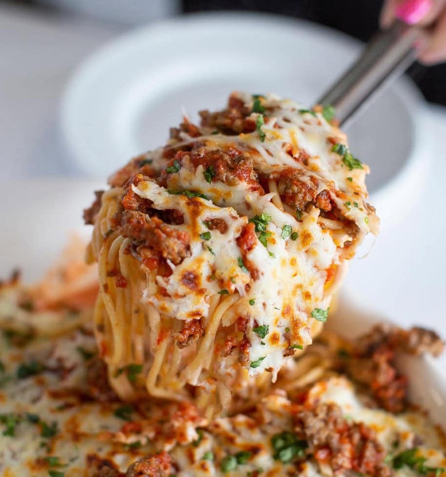A close-up of a serving of cheesy baked spaghetti being lifted with tongs. The spaghetti is covered in melted cheese, tomato sauce, ground meat, and garnished with chopped herbs. A white plate is visible in the background.