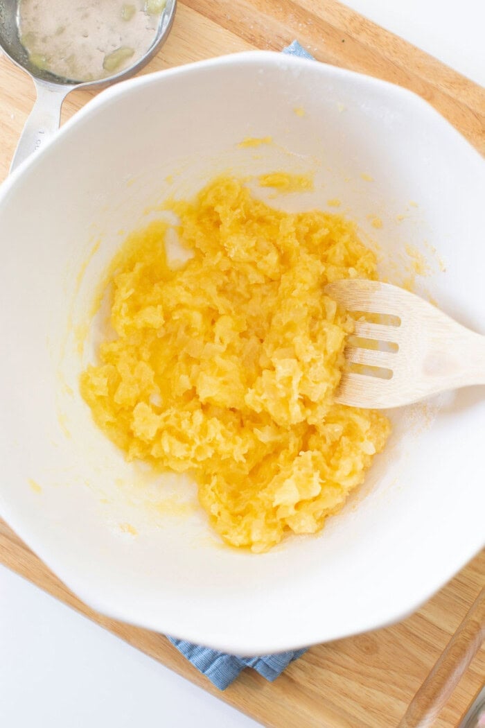 A white bowl filled with mashed yellow substance, possibly potatoes or Easter fluff, sits on a wooden surface. A wooden spoon rests in the bowl while a small pan with liquid is partially visible in the top left corner.