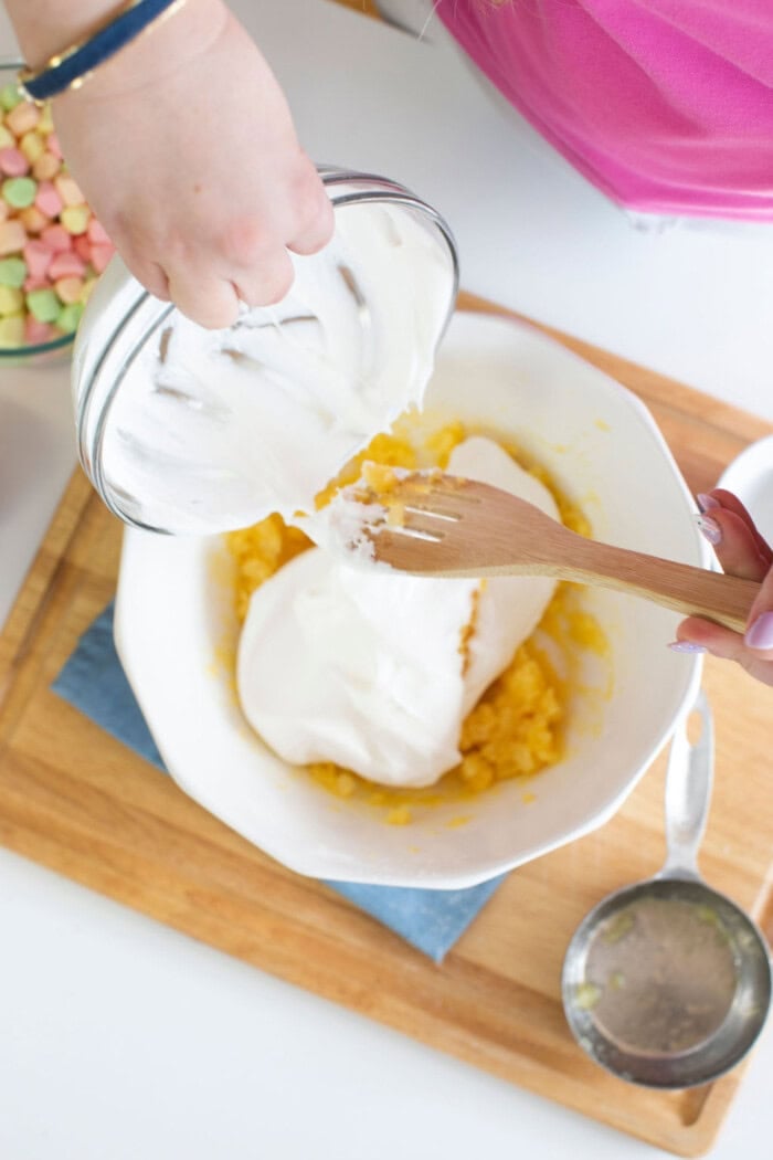 Two hands mix ingredients in a white bowl on a wooden board, crafting an Easter fluff. One hand pours a clear bowl of white cream, while the other stirs with a wooden spoon. Nearby, colorful marshmallows peek out, hinting at the sweet dessert to come.