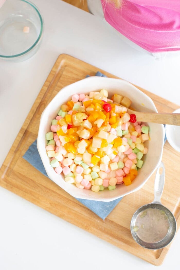 A vibrant fruit salad with pastel marshmallows, canned fruit pieces, and cherries sits charmingly on a wooden cutting board. This delightful Easter fluff is accompanied by a blue napkin, with a spoon and glass bowl completing the picturesque scene.