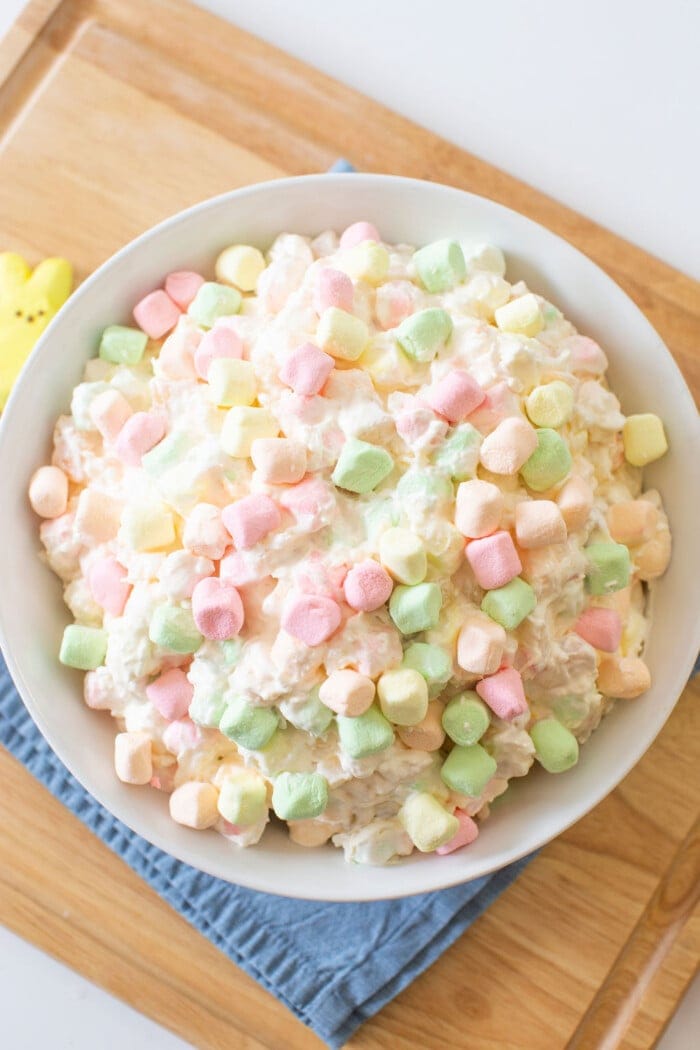 A bowl of ambrosia salad, often dubbed as Easter fluff, rests on a wooden cutting board with a blue cloth underneath. The salad is crowned with pastel-colored mini marshmallows, offering a vibrant and textured display.