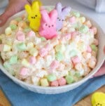 A bowl of pastel-colored Easter fluff salad topped with three bunny-shaped marshmallow candies in yellow, pink, and purple. The fluffy concoction rests on a blue napkin, while a persons hands are partially visible holding the delightful bowl.