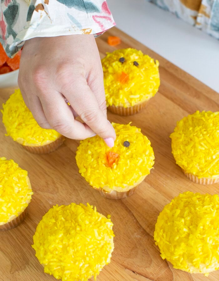 Process photo of making the Easter chick cupcakes face with mini chocolate chips for the 2 eyes and orange candy for the beak.