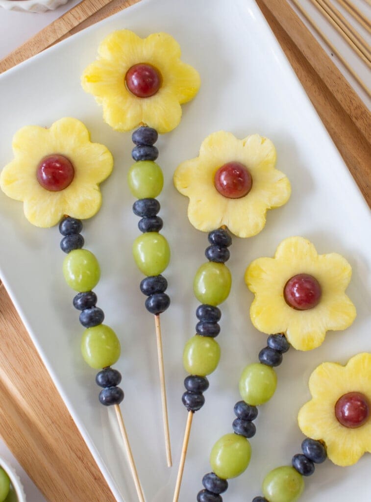 Close-up of fruit skewers: grapes, blueberries, pineapple flowers, and grapes in center, arranged on a white serving tray.