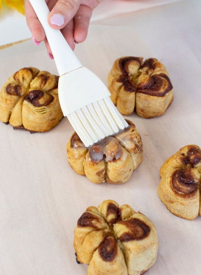 Brushing icing over pumpkin cinnamon rolls on a baking sheet.