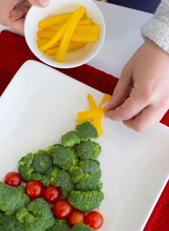 Hands decorate a Christmas veggie tray with broccoli and cherry tomatoes, while a yellow bell pepper star is placed on top. A bowl of pepper strips sits in the background.