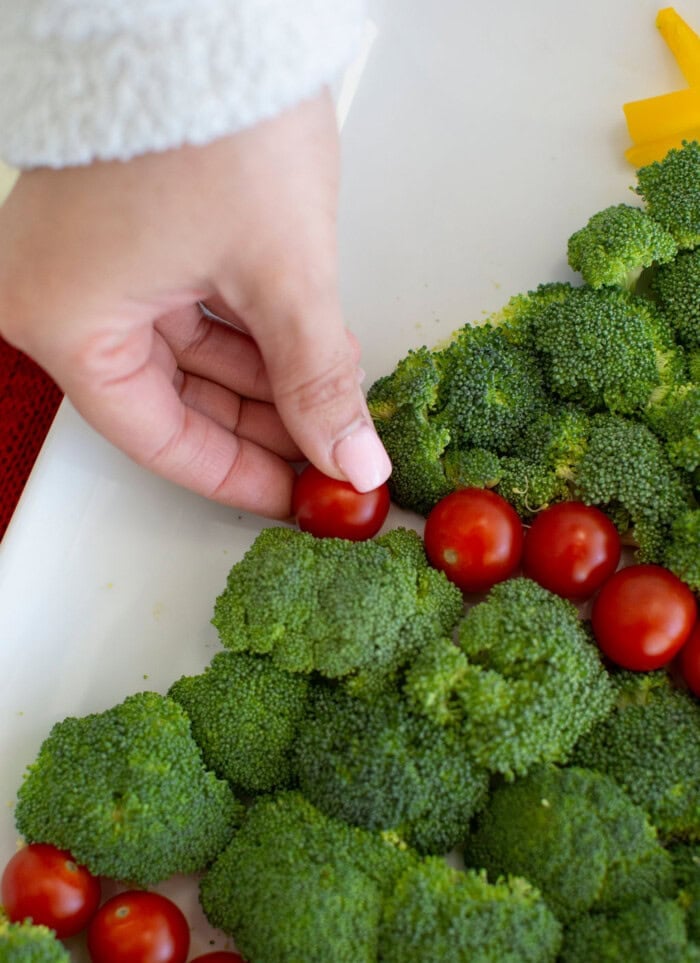 A hand arranges broccoli and cherry tomatoes into a tree shape for a Christmas veggie tray.