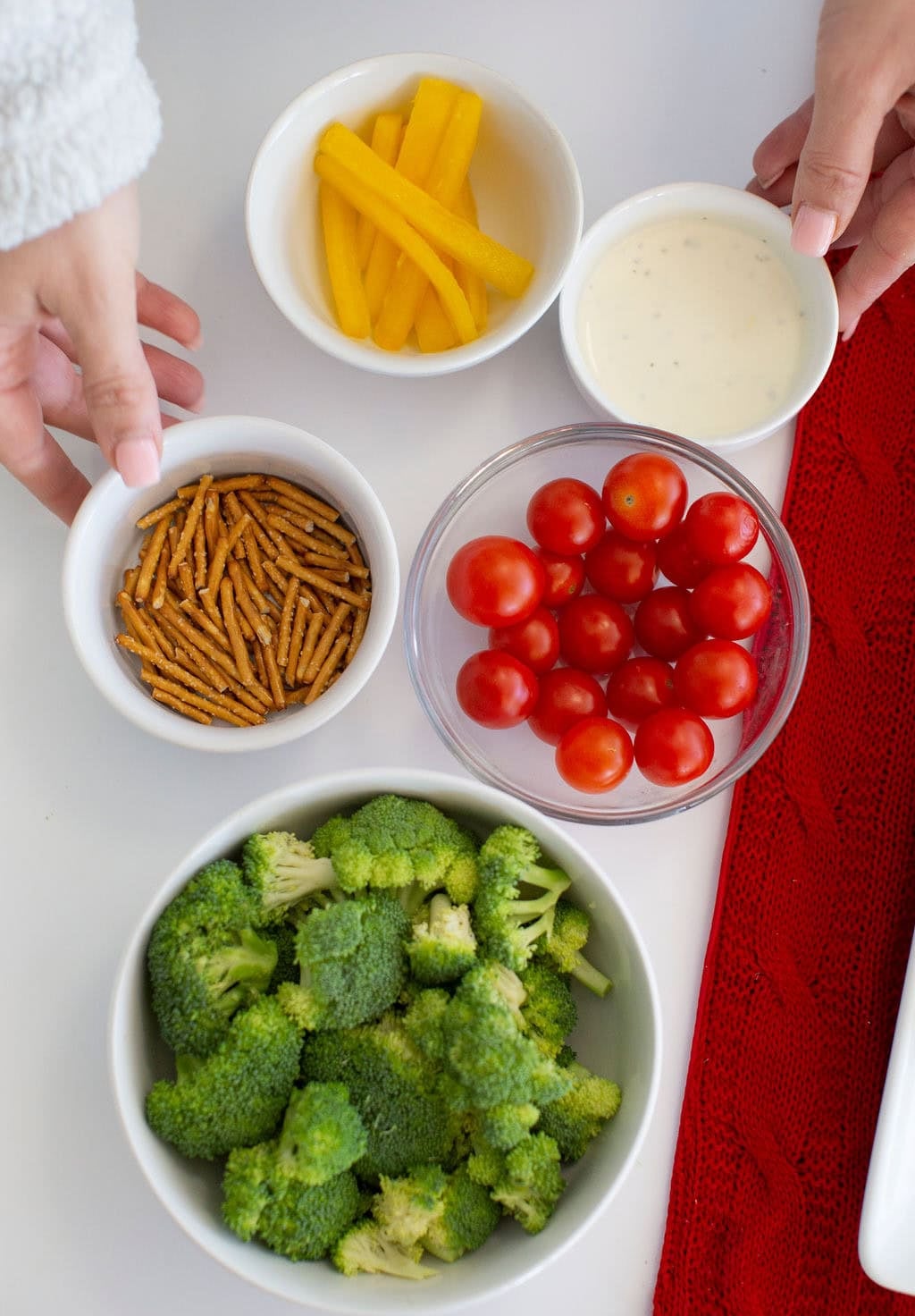 A top-down view of ingredients for a Christmas veggie tray, with bowls of broccoli florets, cherry tomatoes, pretzel sticks, yellow bell pepper slices, and creamy dip being arranged on a white surface.