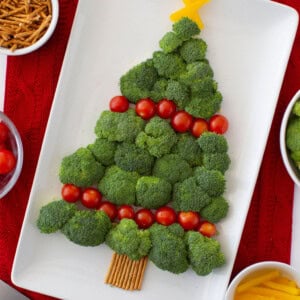 Broccoli arranged in the shape of a Christmas tree on a white platter, decorated with cherry tomatoes and bell pepper strips as ornaments. Pretzel sticks form the trunk. The platter is on a red cloth, surrounded by bowls of snacks.