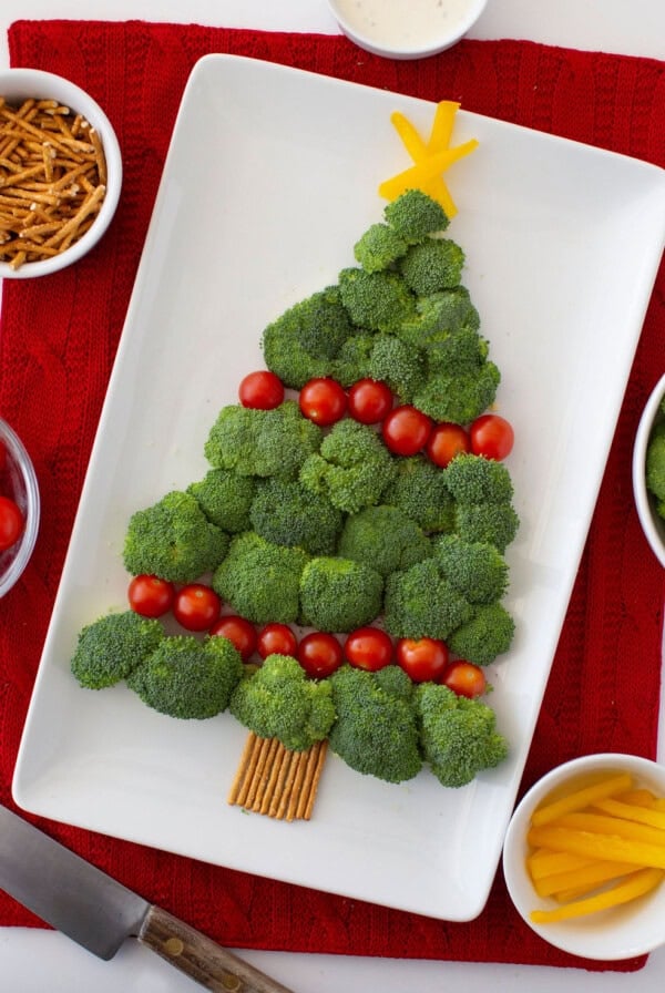 Broccoli arranged in the shape of a Christmas tree on a white platter, decorated with cherry tomatoes and bell pepper strips as ornaments. Pretzel sticks form the trunk. The platter is on a red cloth, surrounded by bowls of snacks.