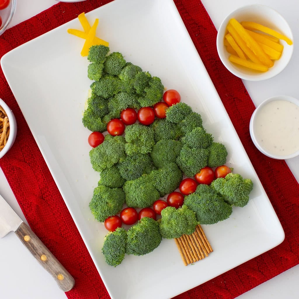 A white rectangular platter displays a festive Christmas veggie tray with broccoli arranged into a Christmas tree, decorated with cherry tomatoes, yellow bell pepper slices, and a pretzel trunk. Bowls of dipping sauce and sliced yellow bell peppers sit nearby.