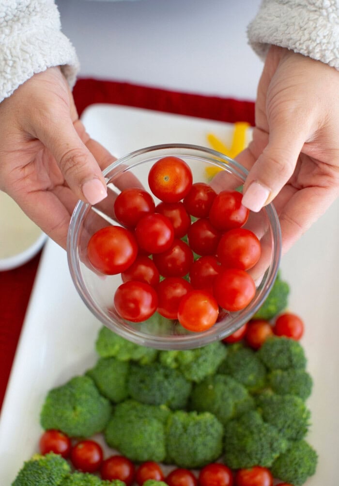 Hands hold a bowl of cherry tomatoes over a platter of broccoli arranged as a Christmas veggie tray, with red pepper slices decorating the tree.