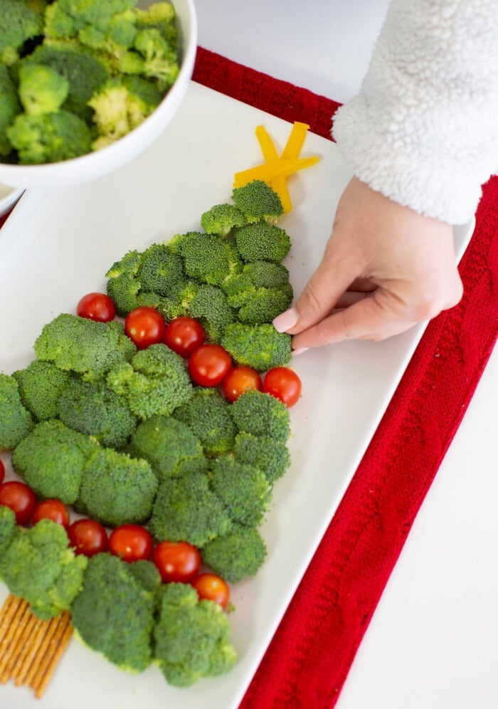 A person arranges cherry tomatoes on a broccoli-shaped Christmas veggie tray, with a cheese star on top and a bowl of broccoli nearby.