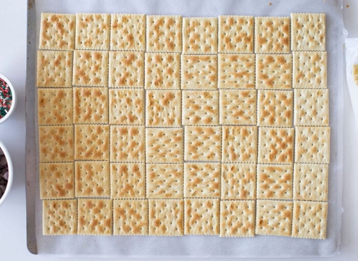 A baking tray lined with parchment paper holds an even grid of saltine crackers. The crackers are golden-brown with a dotted pattern. Small bowls with ingredients are partially visible at the edges.