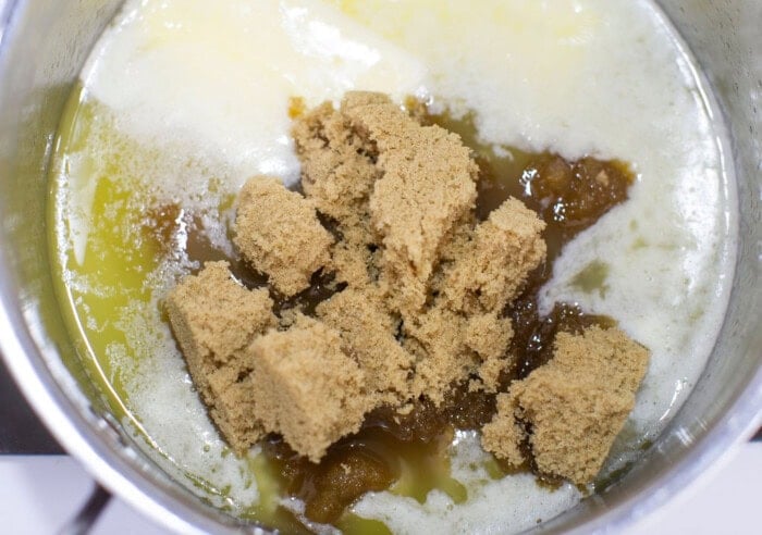A metal pot containing melting butter and brown sugar. The butter is partially melted and pooling around the sugar, creating a mixture of golden liquid and soft, brown chunks ready for blending.