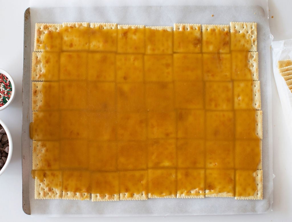 A baking tray topped with a layer of saltine crackers spread with a caramel-like sauce. Small bowls with chocolate chips and festive sprinkles are visible on the left side. Some unspread crackers are on the right.
