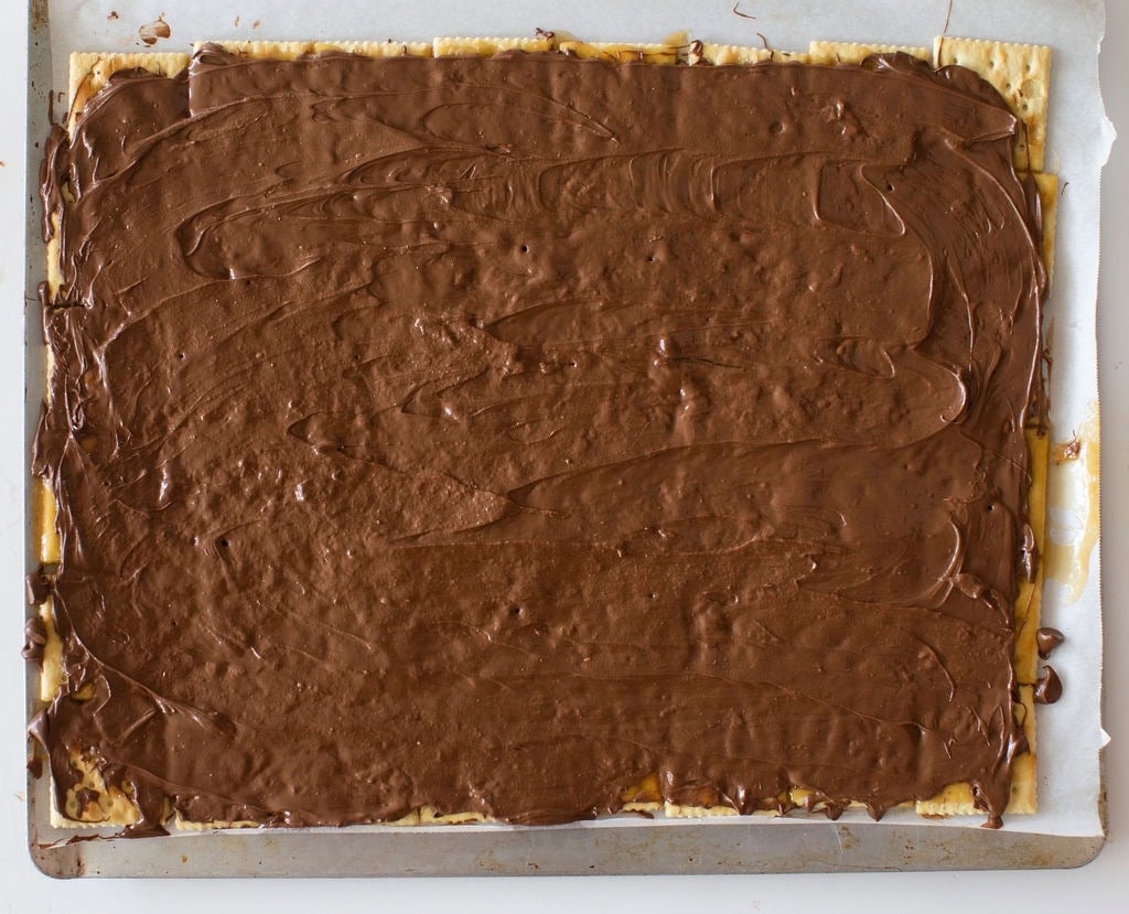 A baking sheet with a layer of matzo crackers covered in a thick, smooth layer of melted chocolate. Some chocolate slightly spills over the edges, and the parchment paper underneath is visible at the corners.