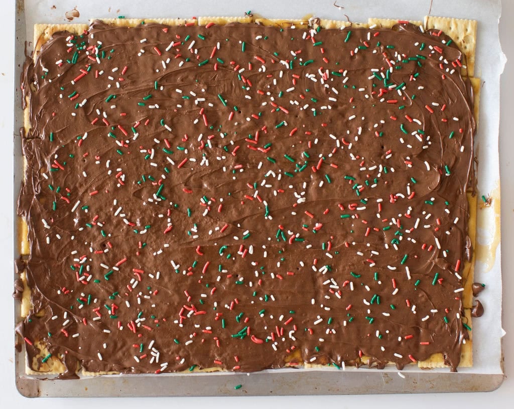 A large baked rectangular pastry topped with a smooth layer of chocolate and scattered red, white, and green sprinkles. Its placed on a metal baking tray lined with parchment paper.
