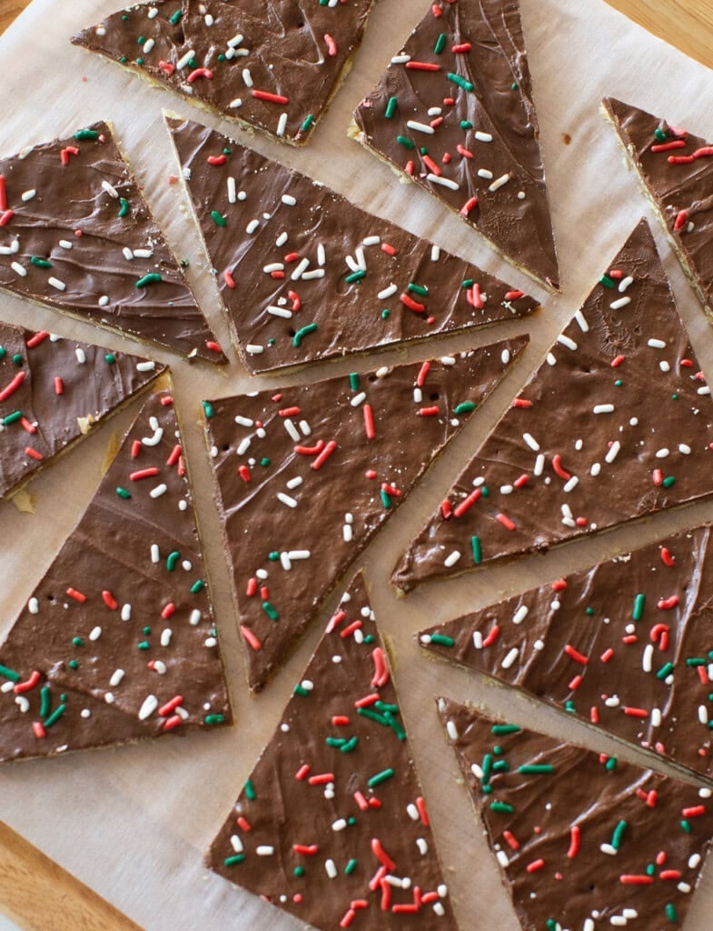 Triangular chocolate bark pieces are topped with festive red, green, and white sprinkles. They are neatly arranged on a parchment-lined surface, ready to be shared and enjoyed.