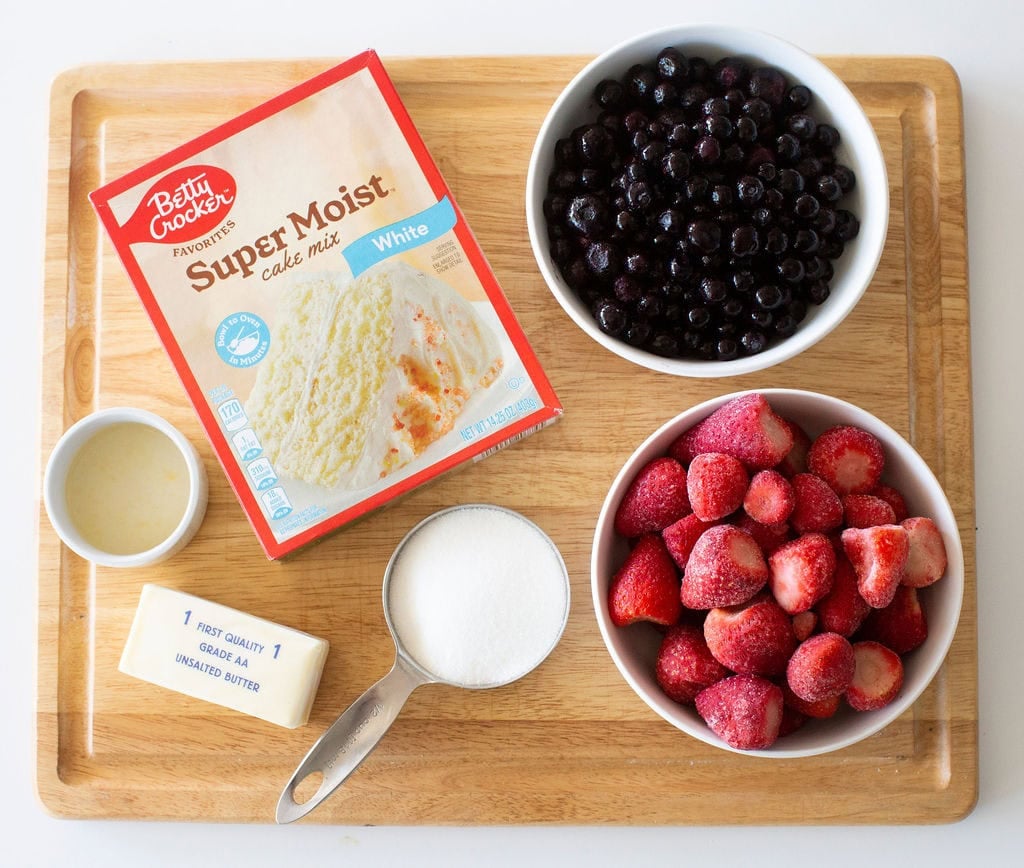 A wooden board displaying ingredients for baking: a box of white cake mix, a bowl of blueberries, a bowl of strawberries, a stick of unsalted butter, a small bowl of liquid, and a measuring cup with sugar.