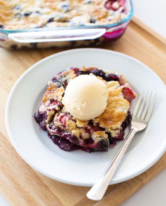 A slice of berry cobbler topped with a scoop of vanilla ice cream is served on a white plate with a fork. The baking dish with the rest of the cobbler is visible in the background on a wooden surface.