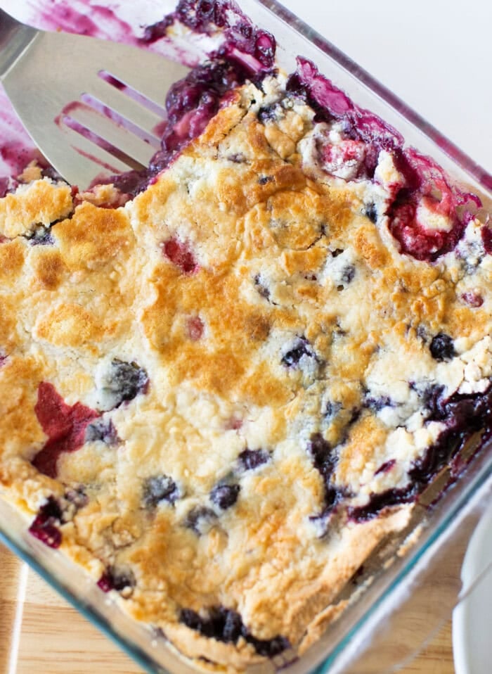 A freshly baked berry cobbler in a glass baking dish, featuring a golden-brown crust with visible blueberries and raspberries peeking through. A metal spatula rests in the dish, ready for serving.