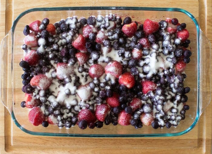A glass baking dish filled with mixed berries, including strawberries, blueberries, and blackberries, topped with granulated sugar, sits on a wooden surface.