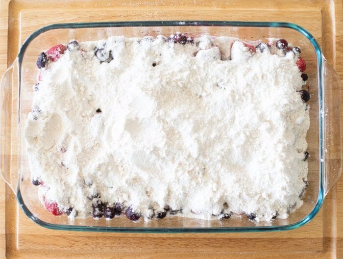 A glass baking dish on a wooden surface filled with a dessert mixture. Visible are mixed berries covered with a layer of flour and sugar, preparing for baking.