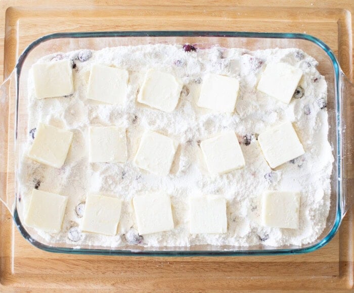 A rectangular glass baking dish filled with a mixture of flour and scattered blueberries. Sixteen evenly spaced, small squares of butter are placed on top. The dish is set on a wooden surface.